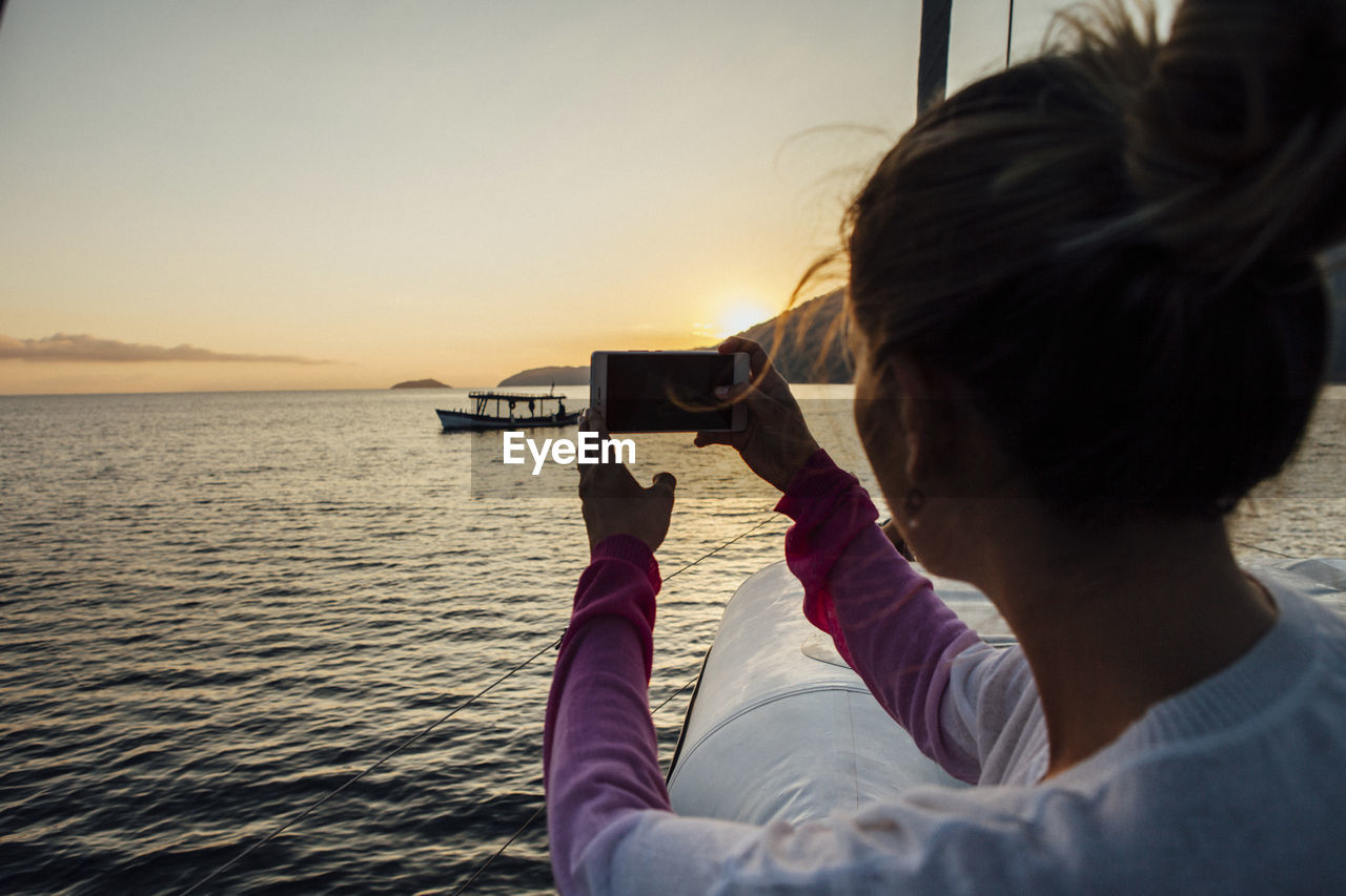 Woman photographing while traveling in sailboat on sea during sunset