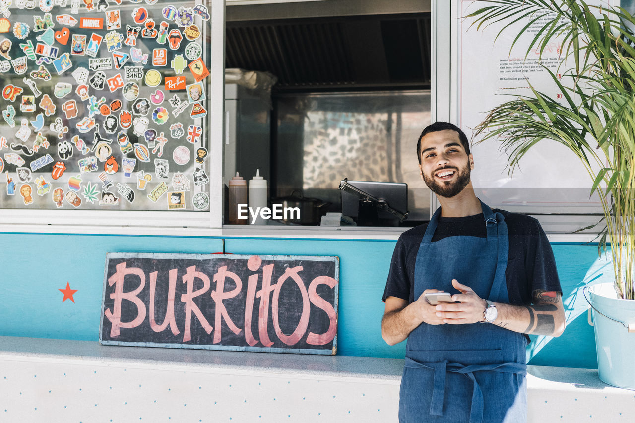 Portrait of smiling young male salesman standing with smart phone against food truck