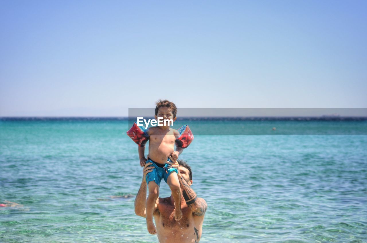 Shirtless father carrying happy son in sea against clear sky