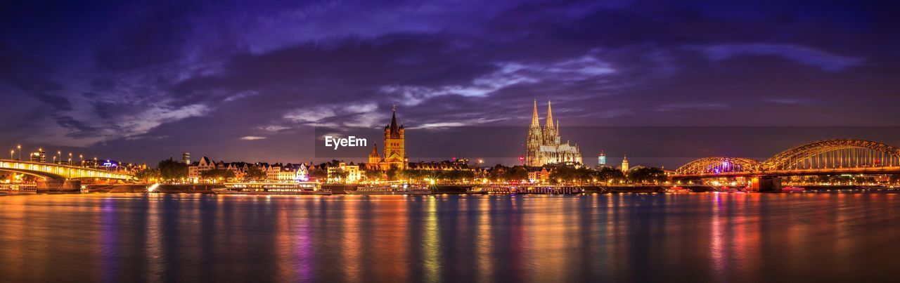 Illuminated buildings by river against cloudy sky