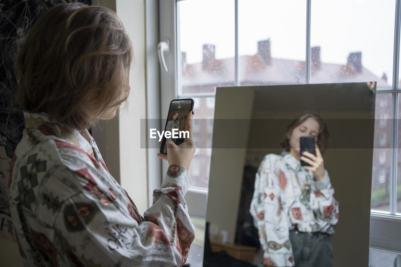 Young woman photographing reflection in mirror through smart phone at home