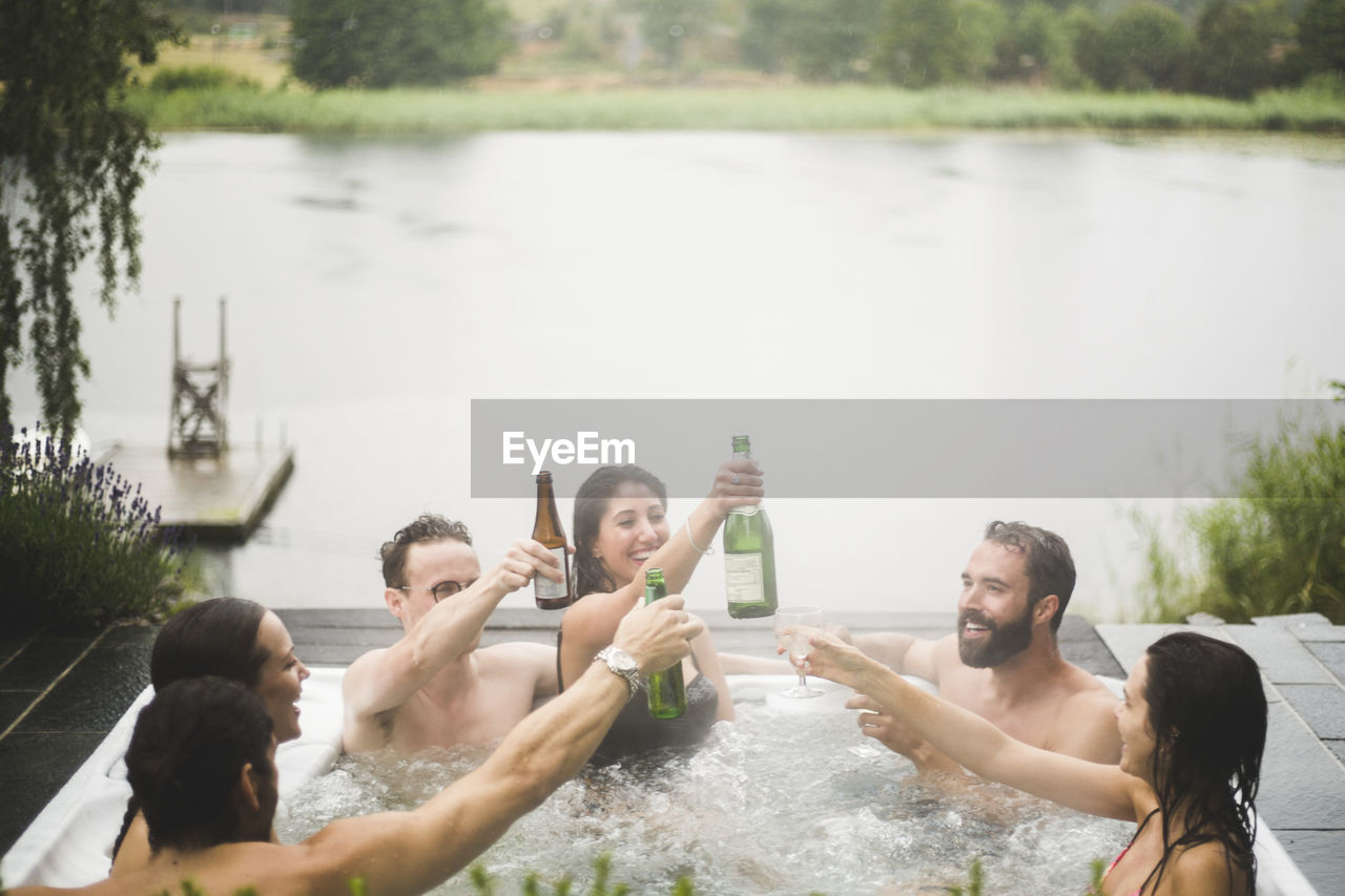 Carefree male and female friends enjoying drinks in hot tub against lake during weekend getaway