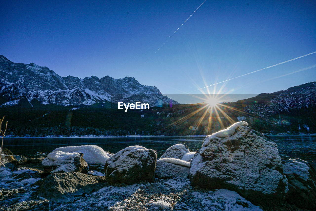sky, mountain, scenics - nature, snow, nature, environment, landscape, winter, cold temperature, beauty in nature, mountain range, blue, sun, tranquility, no people, reflection, rock, sunlight, travel, tranquil scene, star, lens flare, travel destinations, land, clear sky, sunbeam, outdoors, water, cloud, wilderness, snowcapped mountain, morning, non-urban scene, sunrise, mountain peak, dawn, idyllic