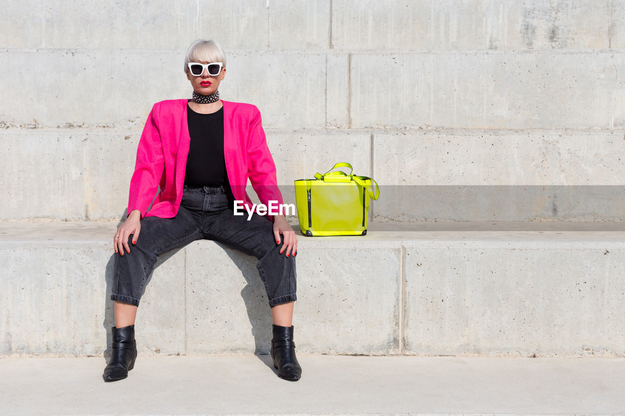 Confident female model wearing bright pink jacket sitting on stairs with vivid yellow handbag in city and looking at camera