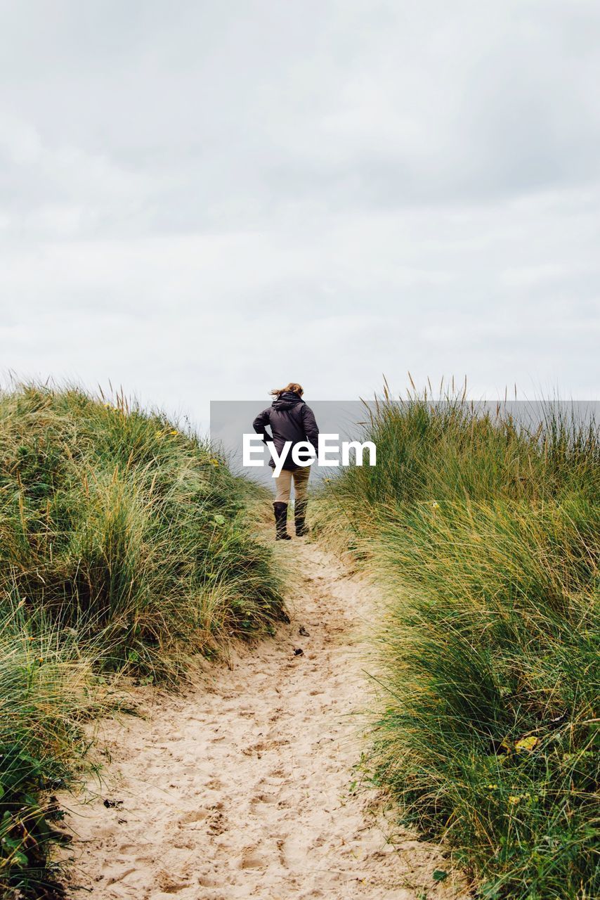 Woman walking on pathway amidst grass against sky