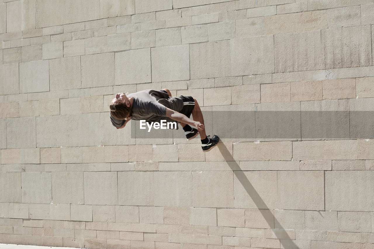 Back view of courageous young male jumping on concrete wall of building while performing dangerous stunt and doing parkour in city