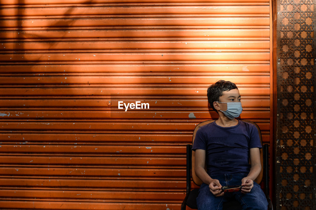 Boy wearing mask looking away while sitting against shutter