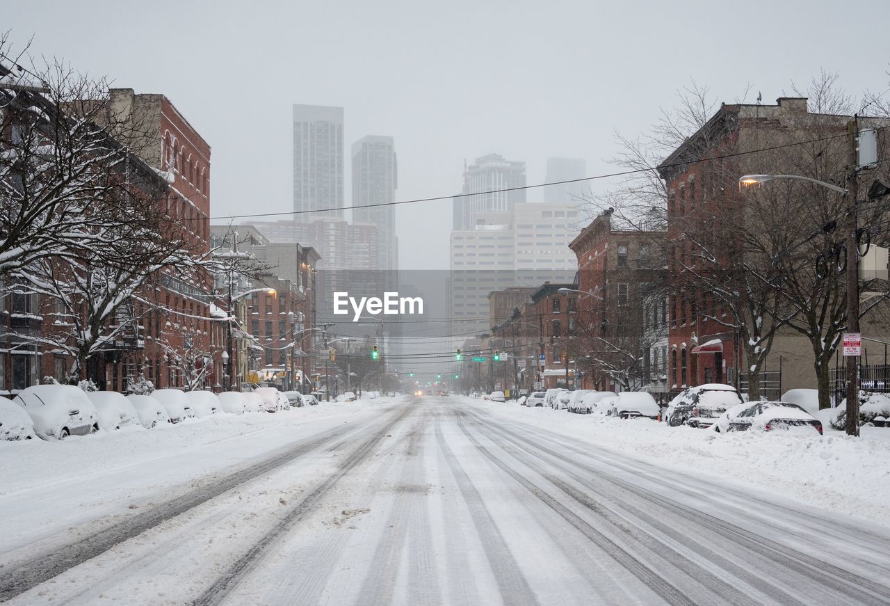 Snow covered road by buildings in city against sky