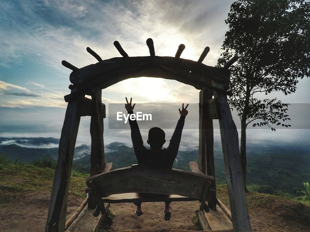 Rear view of man gesturing peace sign on swing against cloudy sky