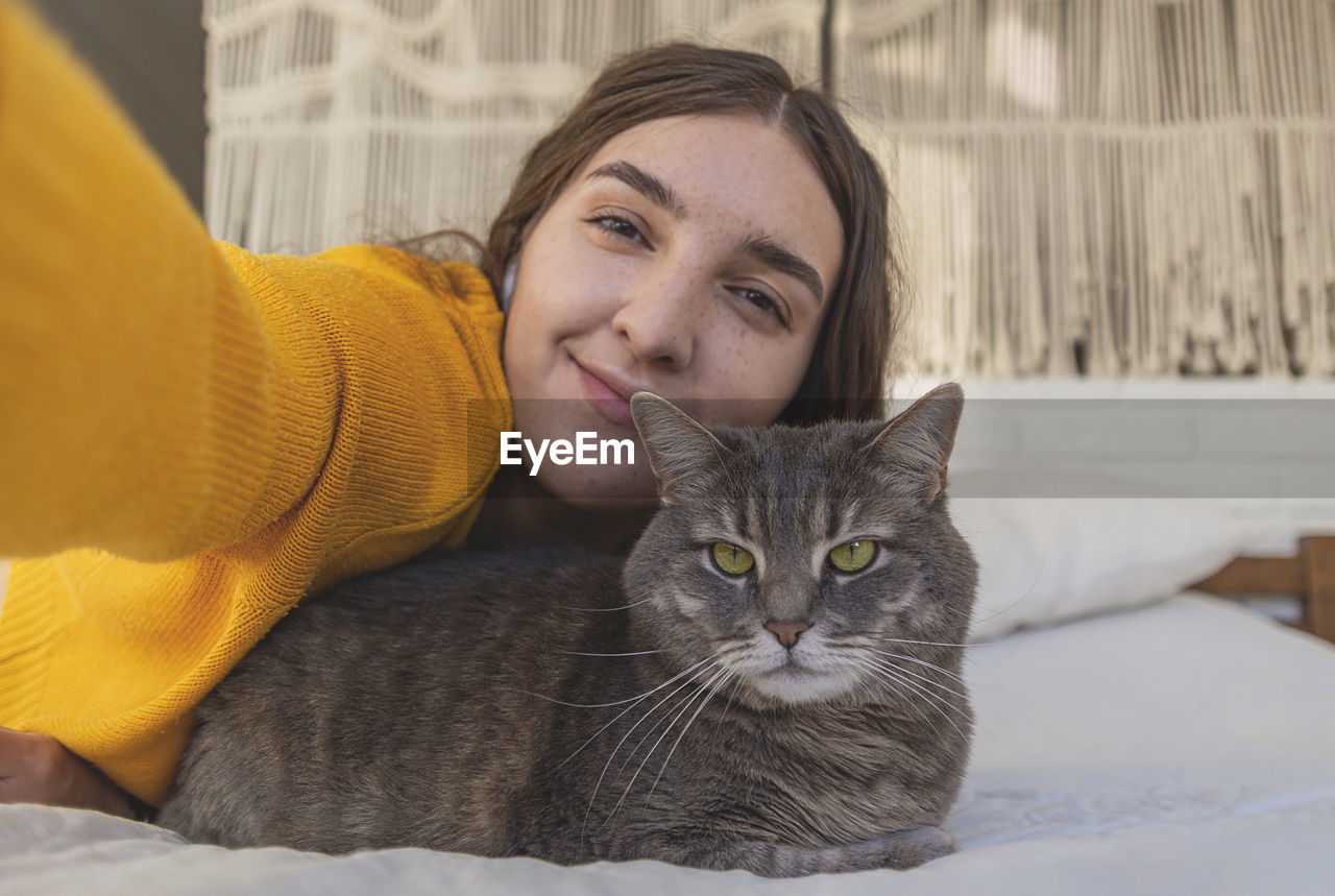 Happy woman in yellow sweater hugs her gray cat on the bed in a light interior