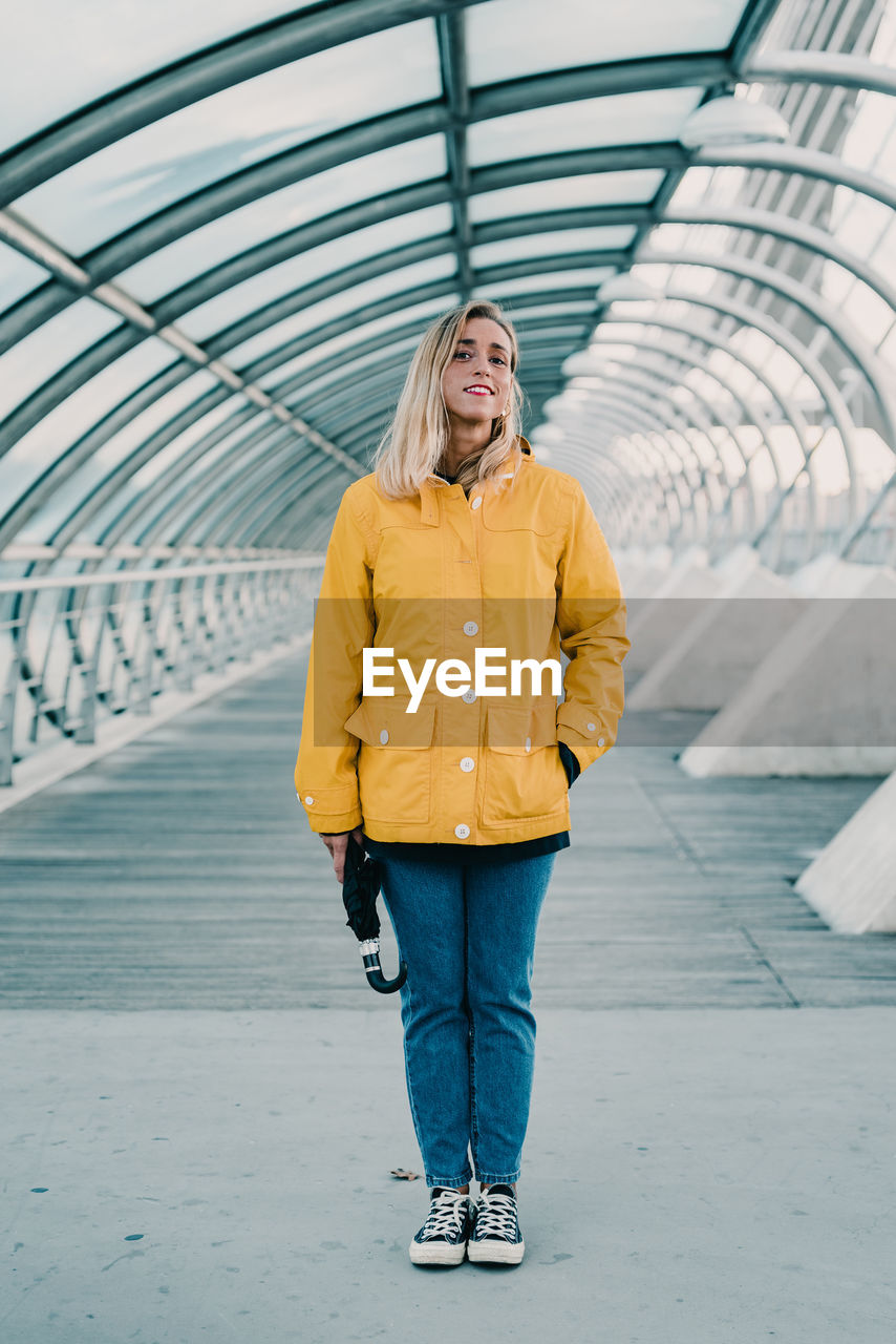 Cheerful female in yellow coat and with umbrella standing under glass roof of tunnel in city and looking at camera