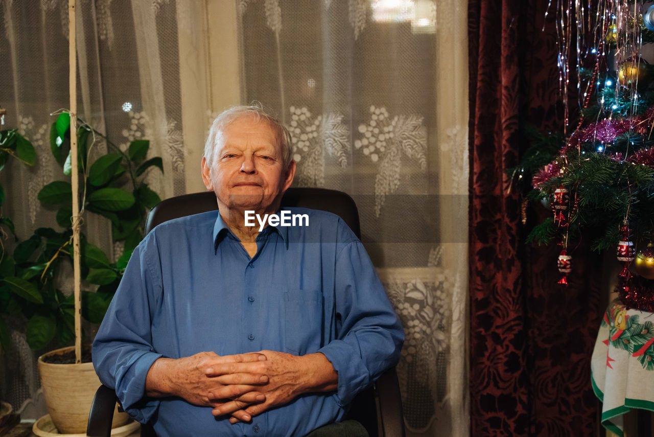 Smiling calm casual senior man in blue shirt spending christmas eve at home and sitting in chair against window next to christmas tree