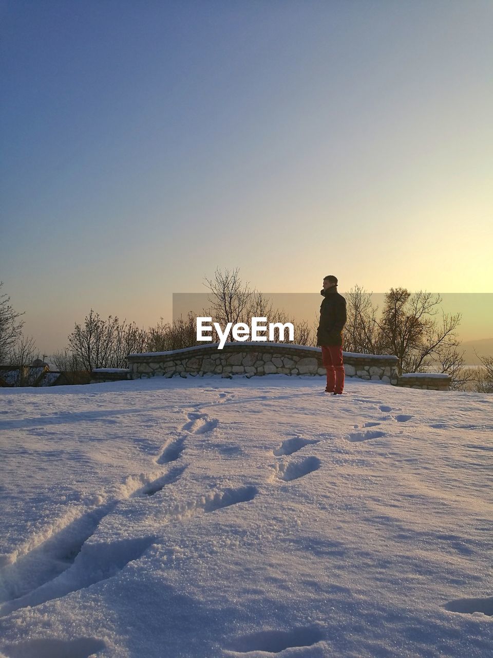 Side view of man standing on snow covered field against sky during sunset