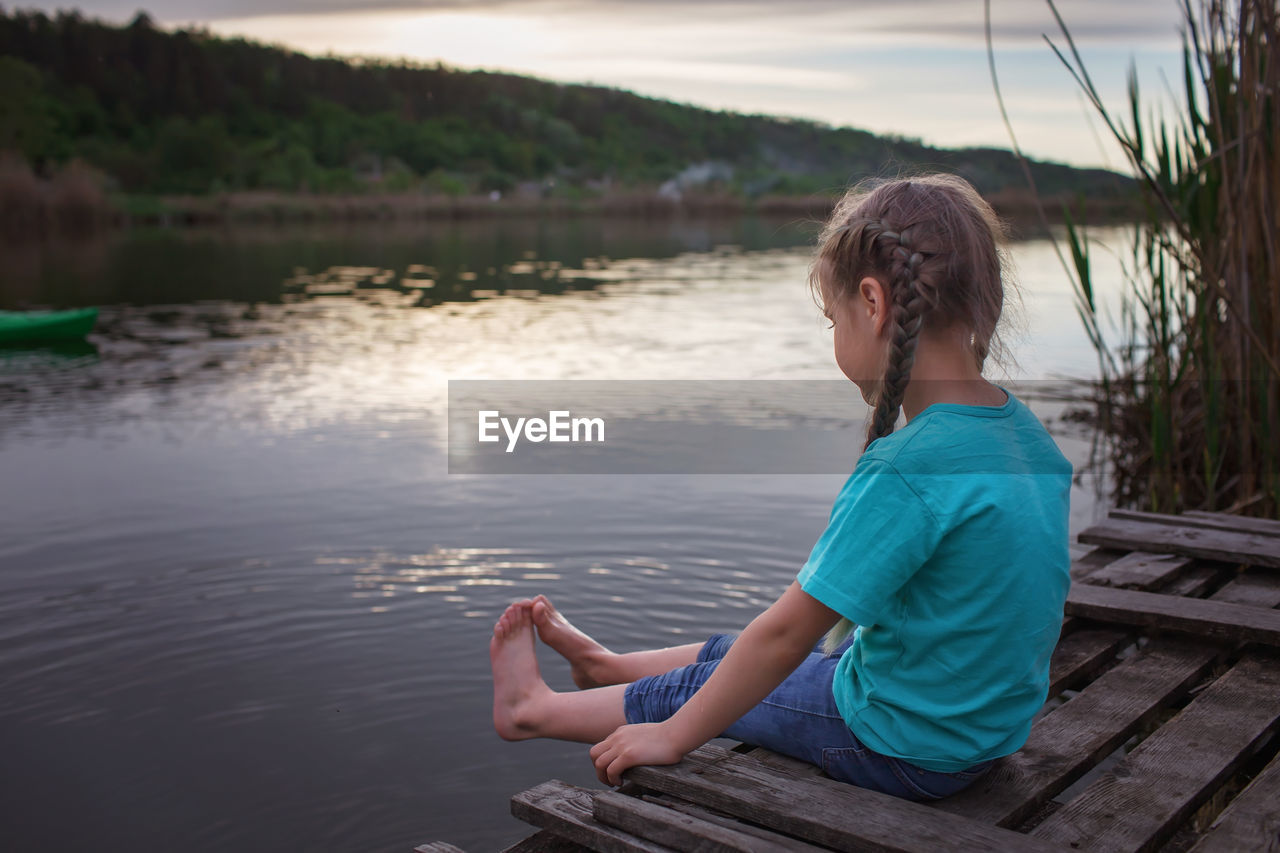 Girl sits on pontoon bridge at lake and enjoys warm sunny evening, happy summertime, countryside