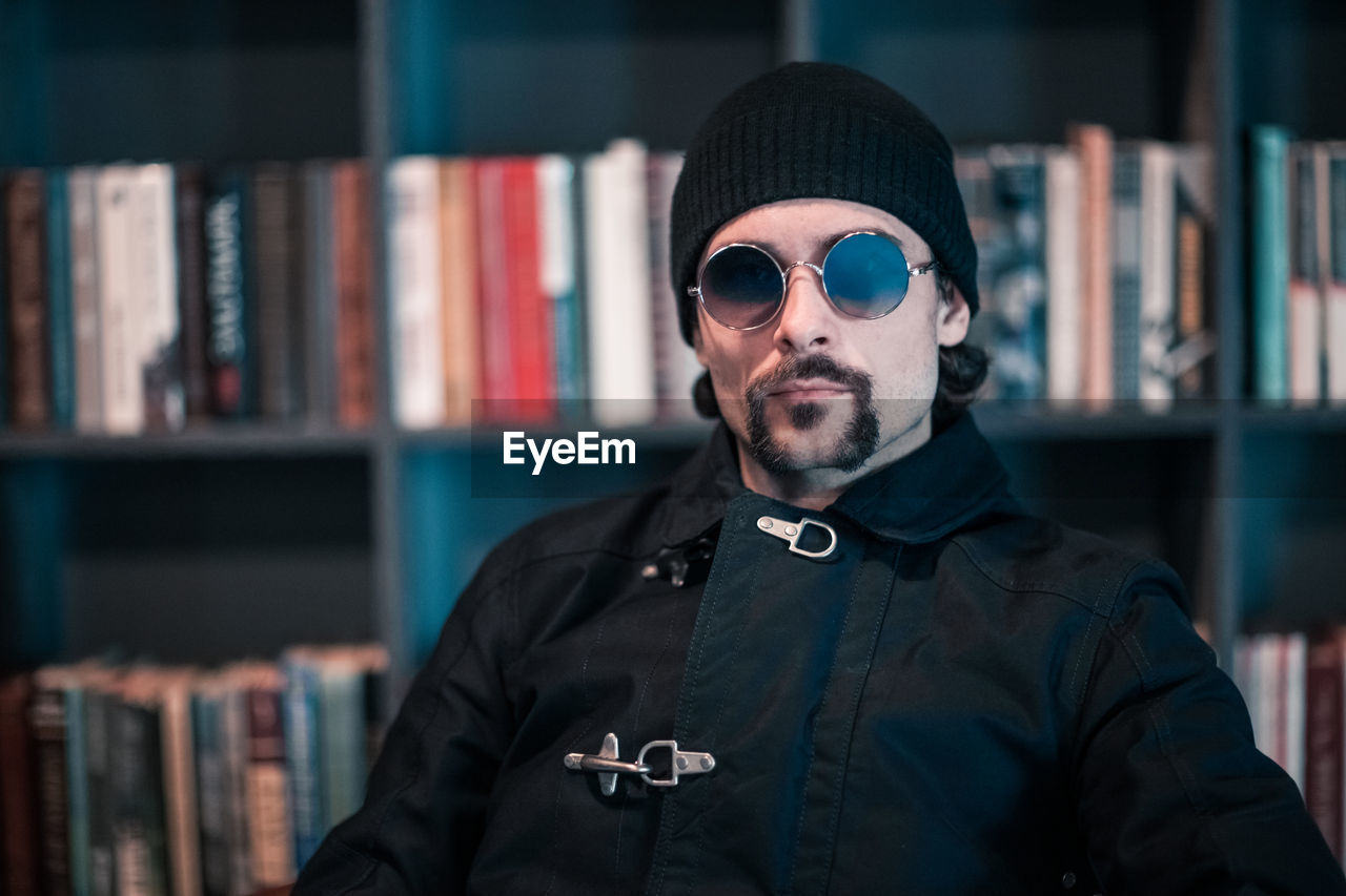 Portrait of handsome man wearing sunglasses while sitting against bookshelf