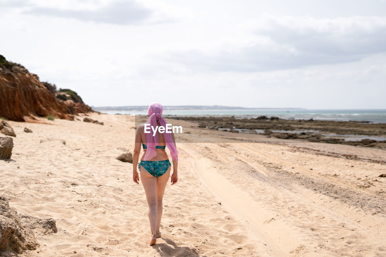 Woman with pink scarf for cancer walking in bikini on the beach