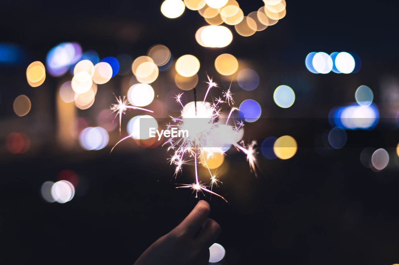 Cropped hand of person holding illuminated sparkler against lights at night