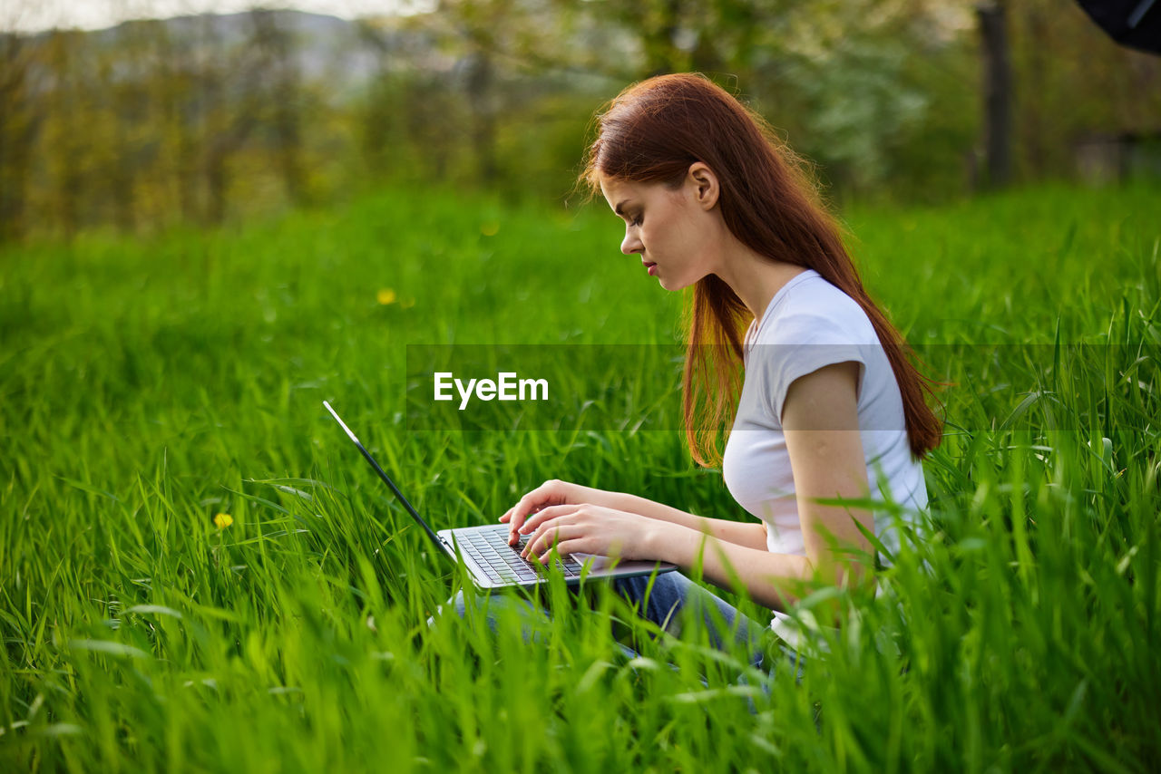 young woman using laptop while sitting on grassy field