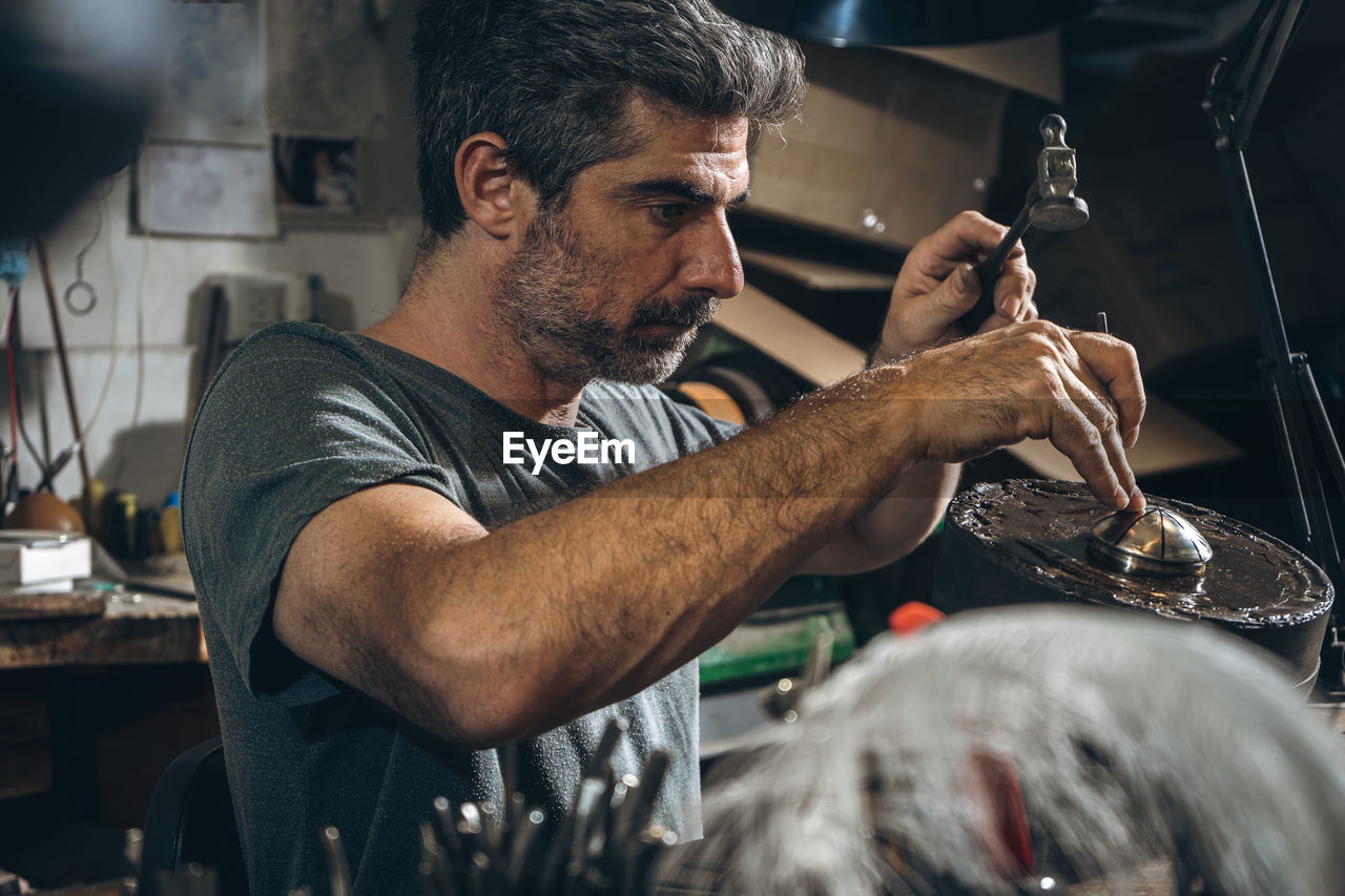 Side view of mature male artisan using metal punch and hammer to carve ornament on silver lid while sitting at table in professional workshop