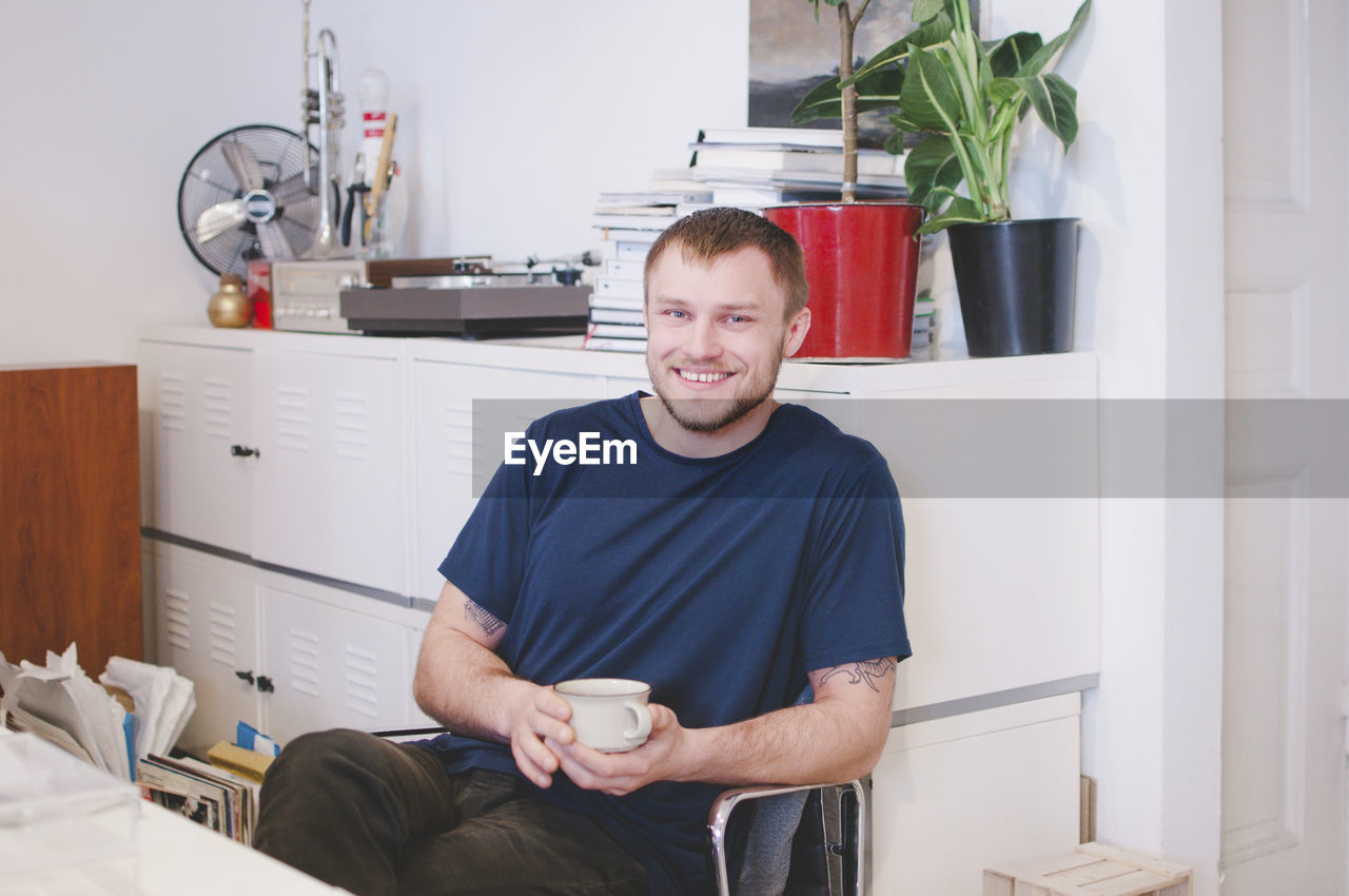 Portrait of happy businessman holding coffee cup while sitting in creative office