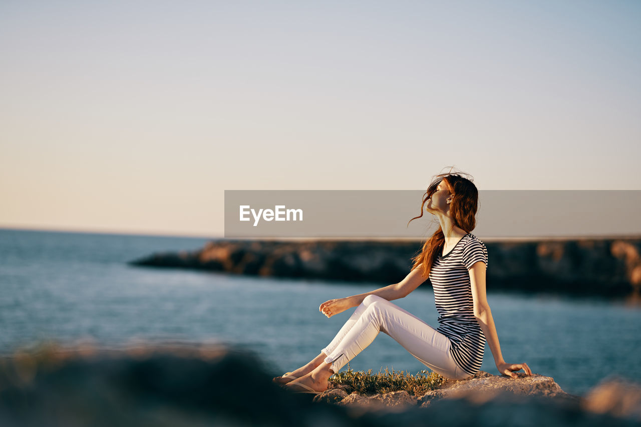 WOMAN SITTING IN SEA AGAINST CLEAR SKY