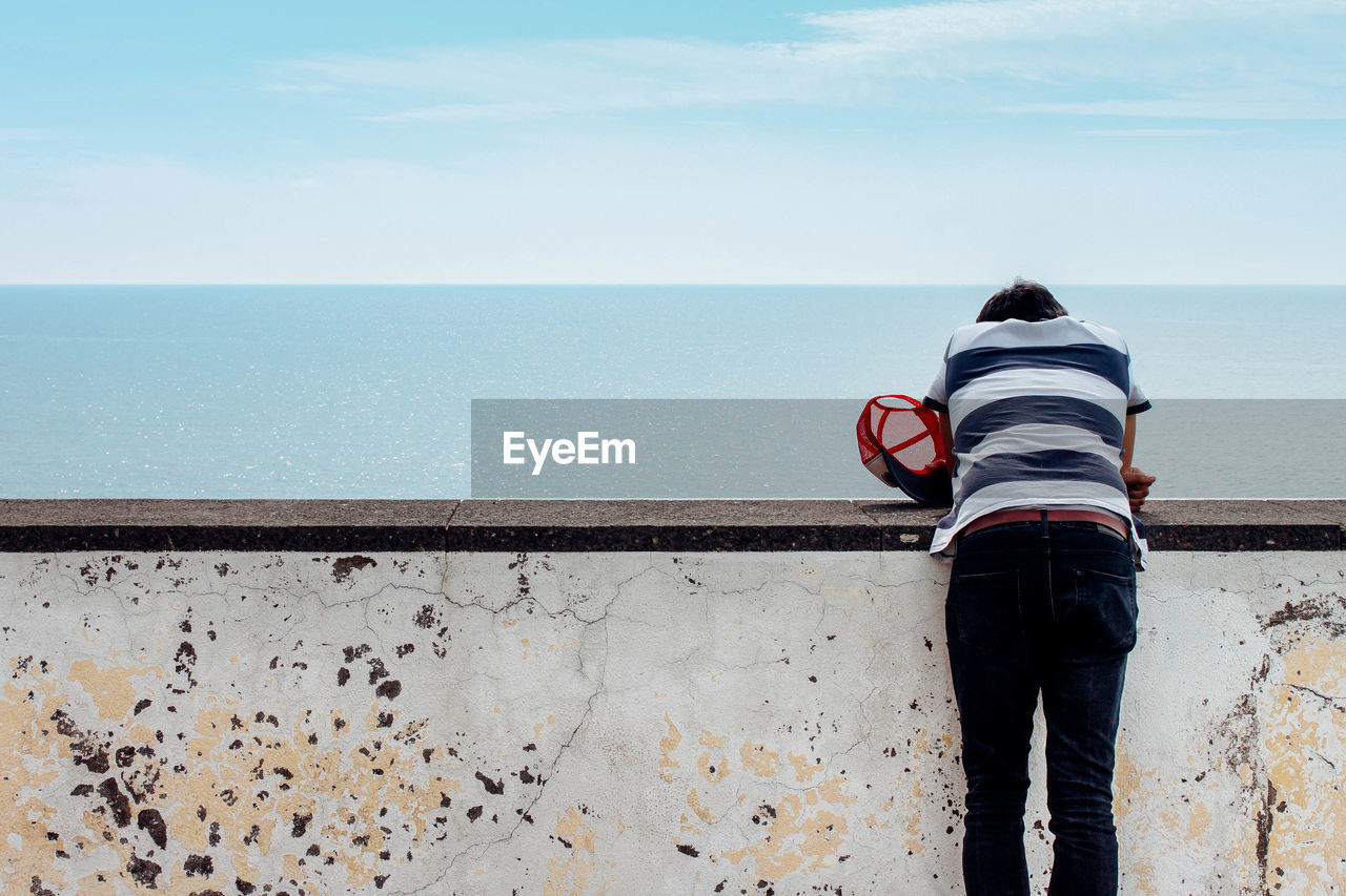 Rear view of person standing at retaining wall by sea against sky