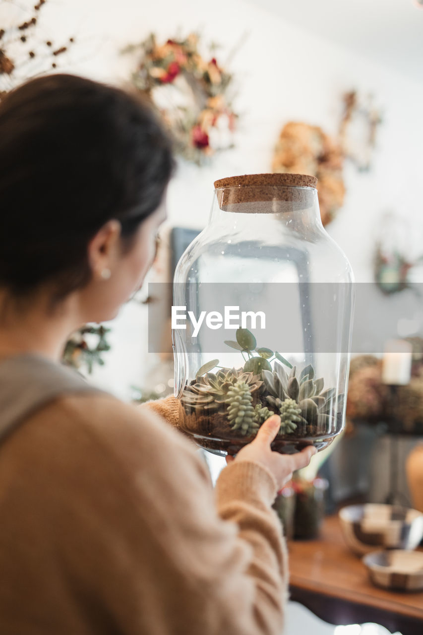 Back view of unrecognizable female in casual clothes holding big transparent glass terrarium closed with cork lid and looking at small green succulents inside while standing against blurred interior of modern flower shop