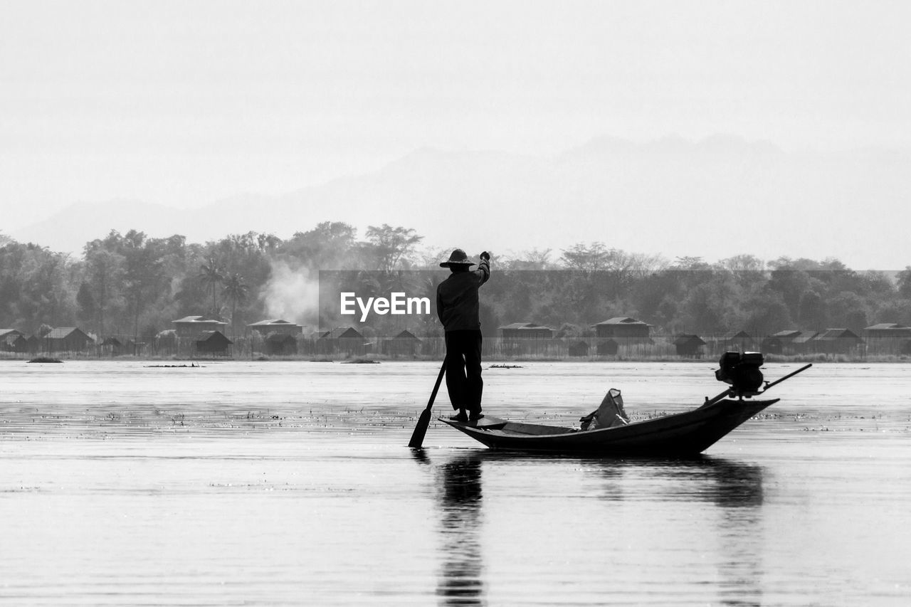 SILHOUETTE MAN STANDING ON BOAT IN LAKE AGAINST SKY