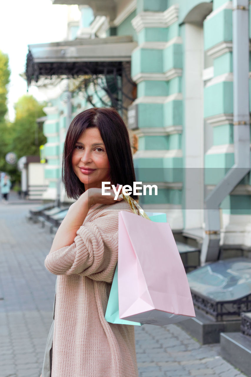 shopping bag, women, shopping, city, one person, smiling, adult, bag, happiness, architecture, consumerism, young adult, retail, street, hairstyle, city life, long hair, cheerful, clothing, emotion, lifestyles, holding, fashion, portrait, customer, standing, casual clothing, building exterior, pink, spending money, store, brown hair, female, carrying, day, looking, looking at camera, outdoors, dress, walking, enjoyment, built structure, city street, focus on foreground, leisure activity, black hair, snapshot, footpath, sidewalk, buying, communication