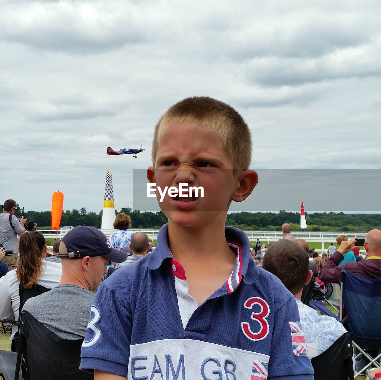 Boy making face while looking away against cloudy sky