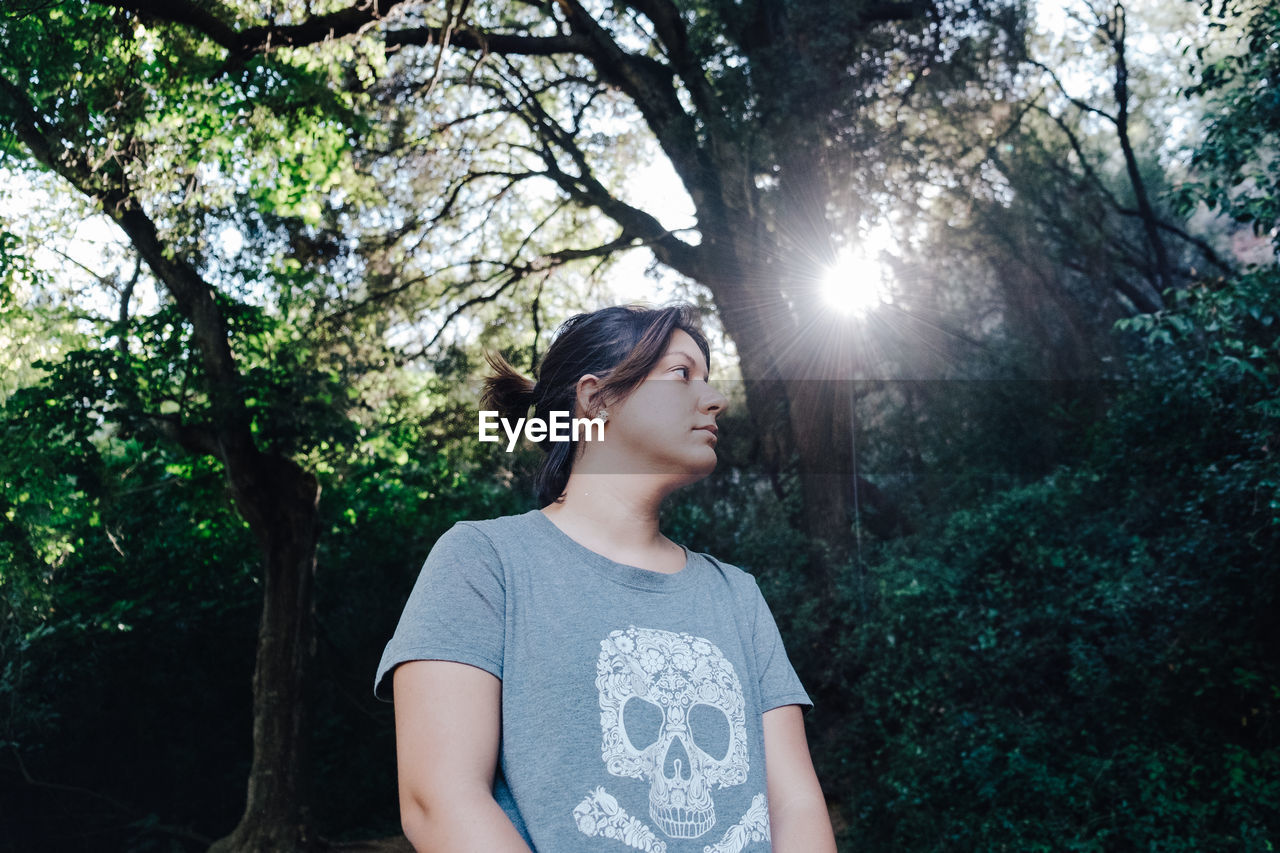 Low angle view of young woman standing against trees in forest on sunny day