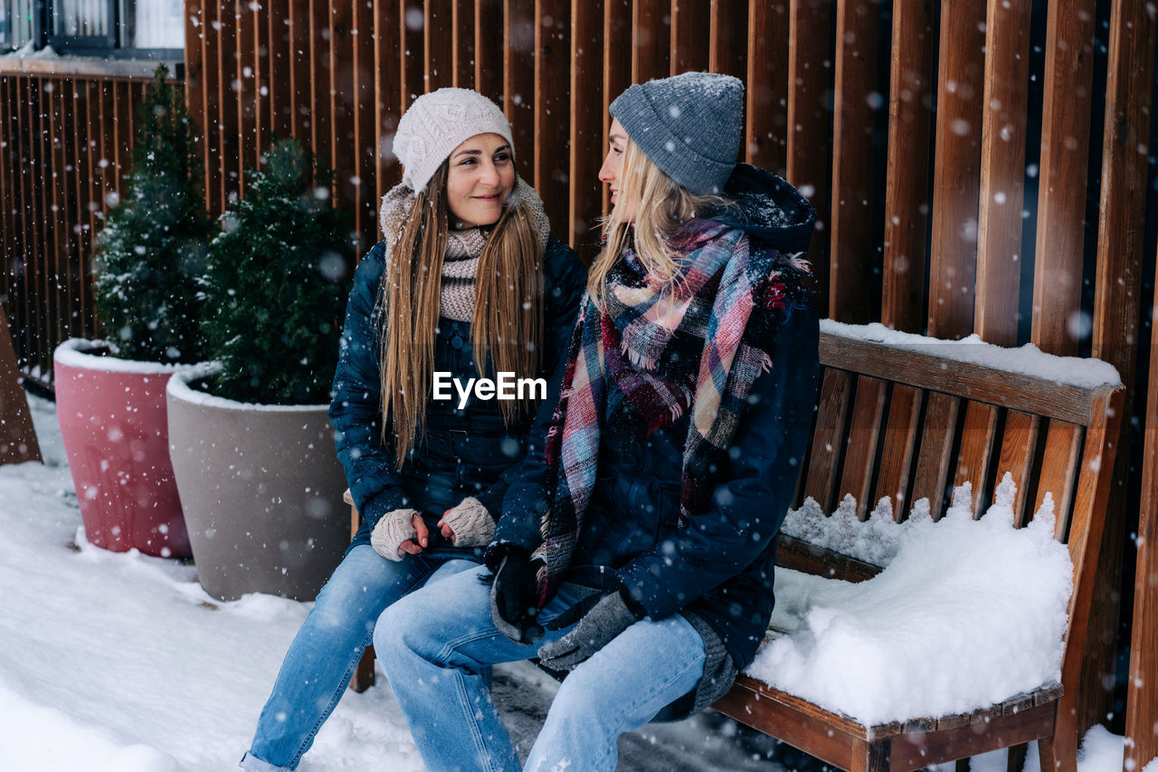 Two women friends in winter coats and hats sitting on a bench on the terrace look at each other.