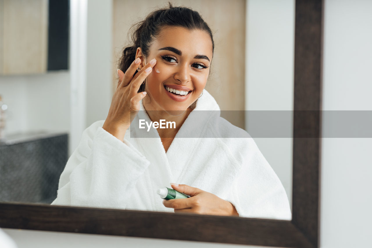 Smiling young woman applying cream on face while looking in mirror