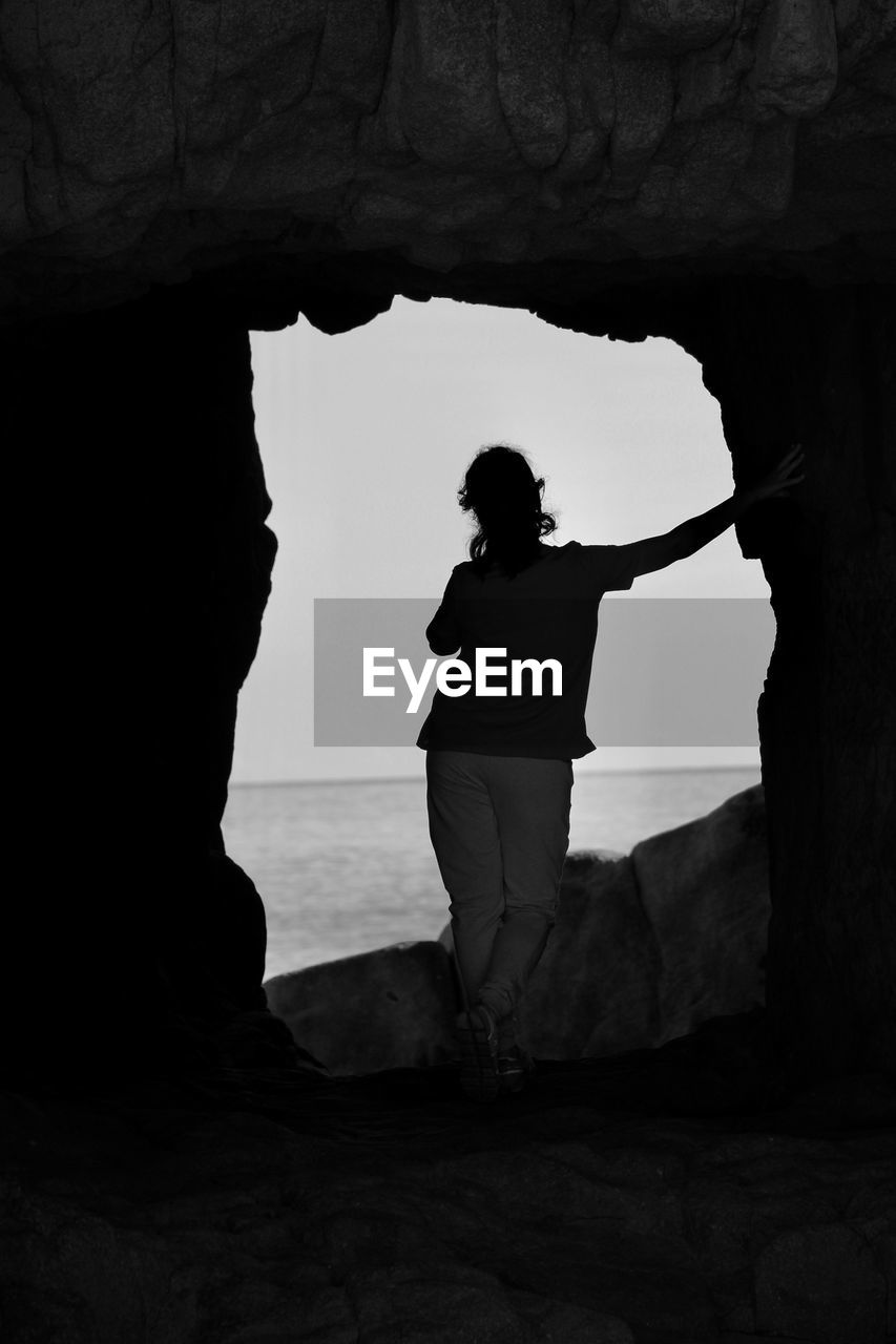 Silhouette of a young woman standing on a hole in the rock against the sky during sunset