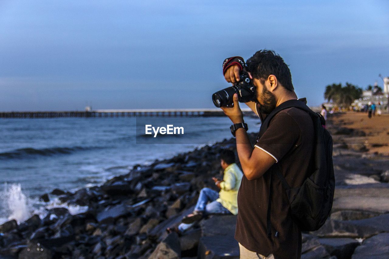 Side view of man on rocky shore photographing sea