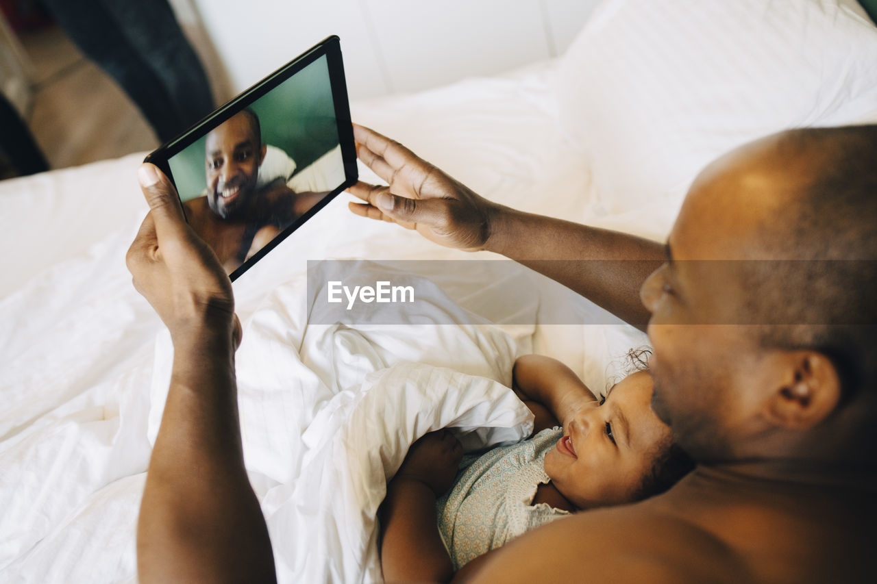 Smiling father and daughter taking selfie on digital tablet while lying in bed at home