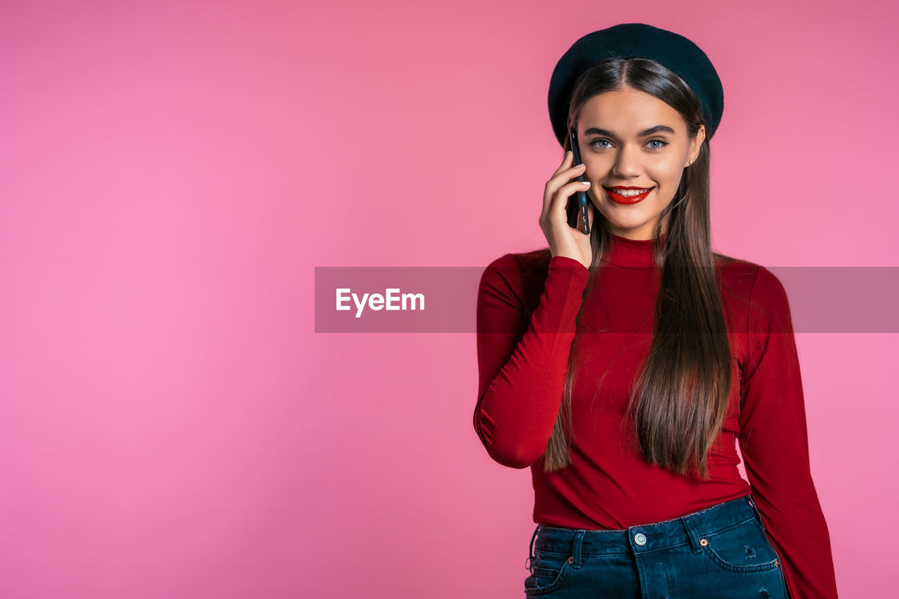 SMILING YOUNG WOMAN USING PHONE WHILE STANDING ON RED WALL