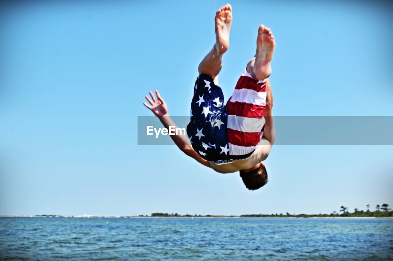 Full length of shirtless man backflipping at beach against clear sky