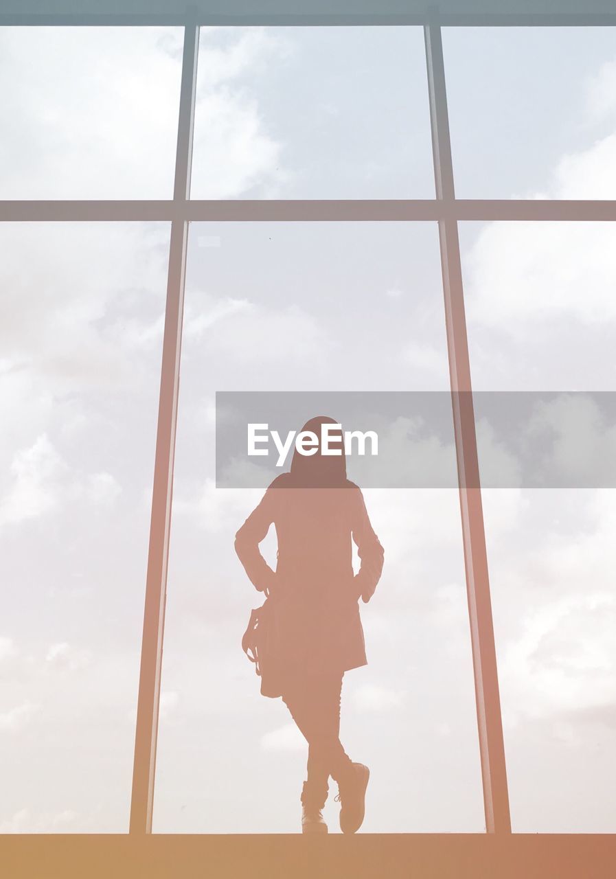 Silhouette woman standing by glass window against cloudy sky