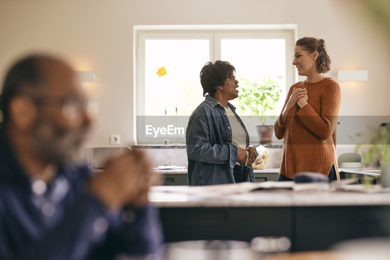 Woman with hands on chest talking to mother-in-law standing in kitchen at home