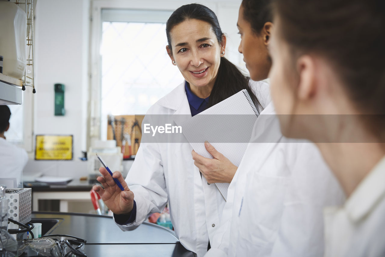 Confident mature teacher explaining to young female college students in chemistry laboratory