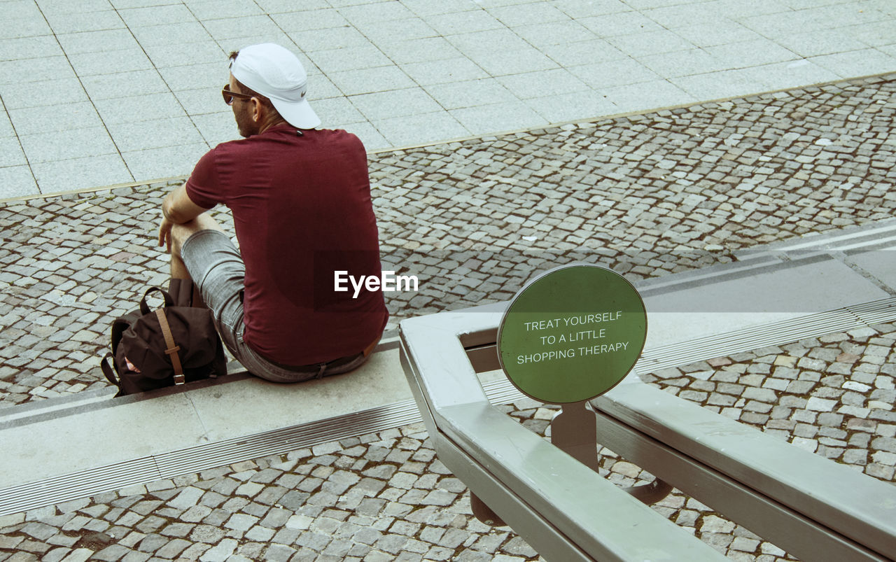 one person, adult, city, communication, rear view, full length, footpath, sitting, flooring, architecture, person, street, men, text, day, cobblestone, hat, clothing, lifestyles, red, high angle view, casual clothing, sign, transportation, floor, outdoors, stone