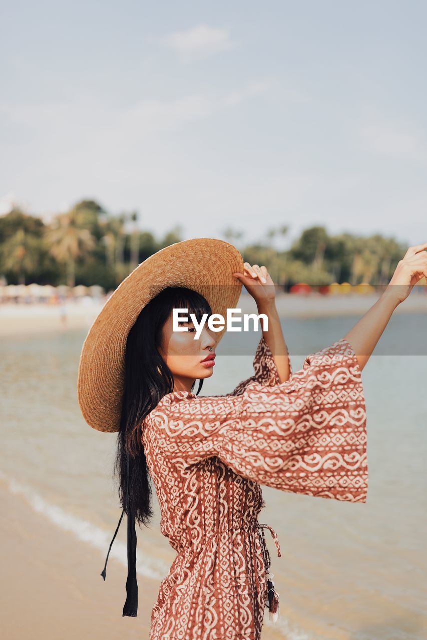 Young woman wearing hat looking away standing at beach against sky