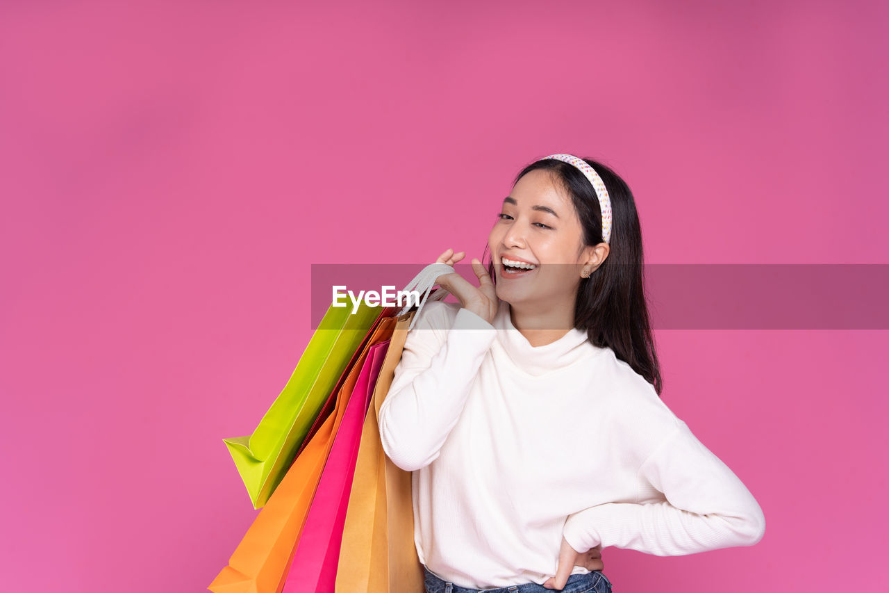 happiness, smiling, pink, women, shopping bag, one person, adult, emotion, cheerful, shopping, colored background, young adult, studio shot, bag, clothing, consumerism, portrait, lifestyles, laughing, fun, long hair, positive emotion, retail, teeth, indoors, smile, enjoyment, casual clothing, copy space, photo shoot, spending money, hairstyle, fashion, holding, fashion accessory, female, pink background, waist up, celebration, customer, looking, standing, joy, person, front view, carrying, outerwear, looking at camera, yellow
