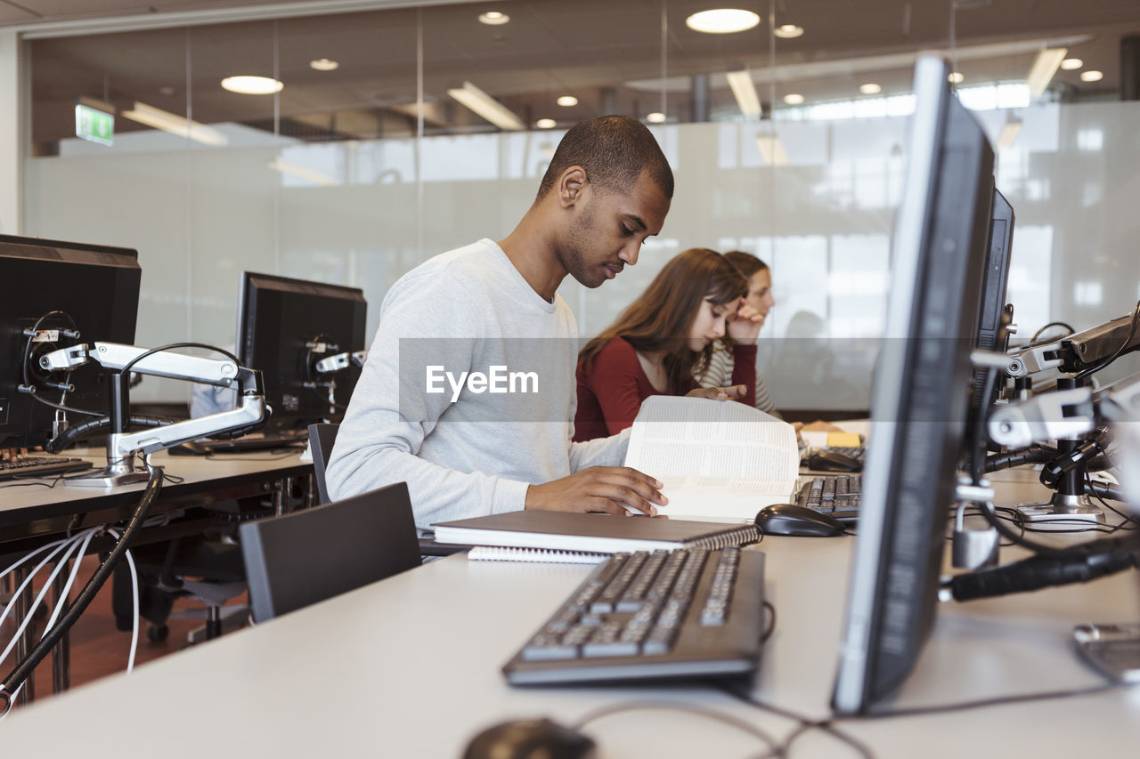 Serious male student reading book while sitting with friends at computer desk in library