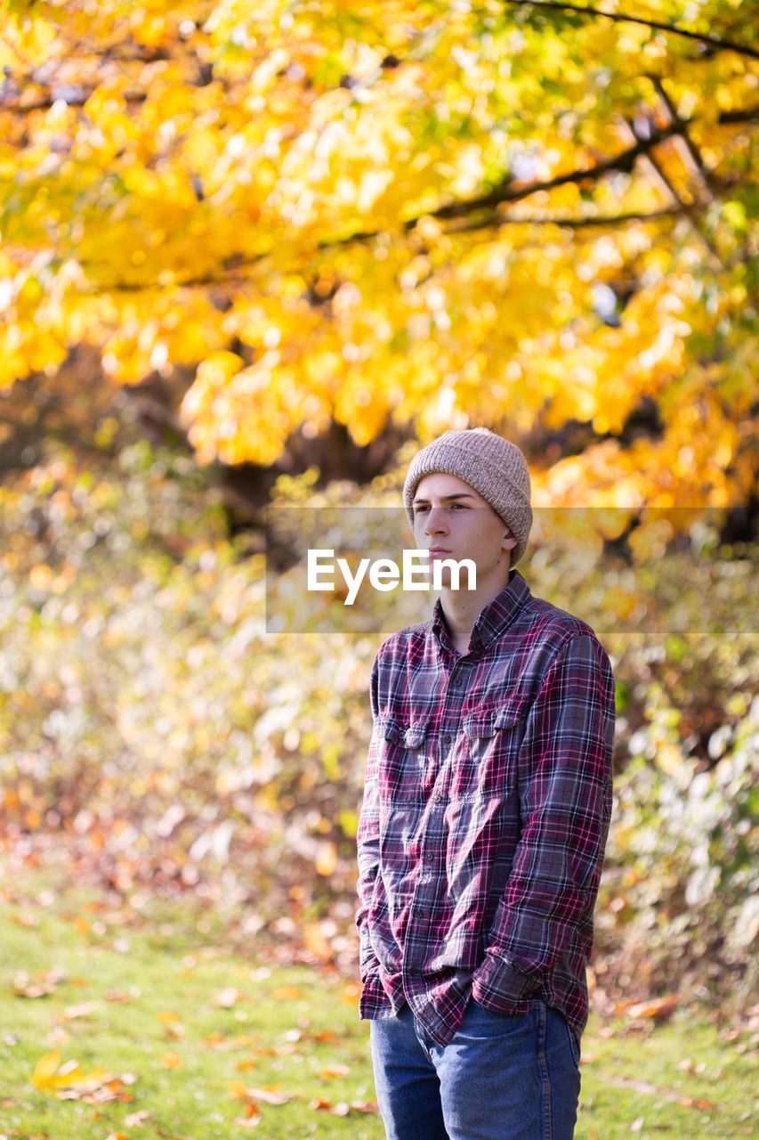 Pensive teen boy in toque and flannel shirt in front of yellow trees