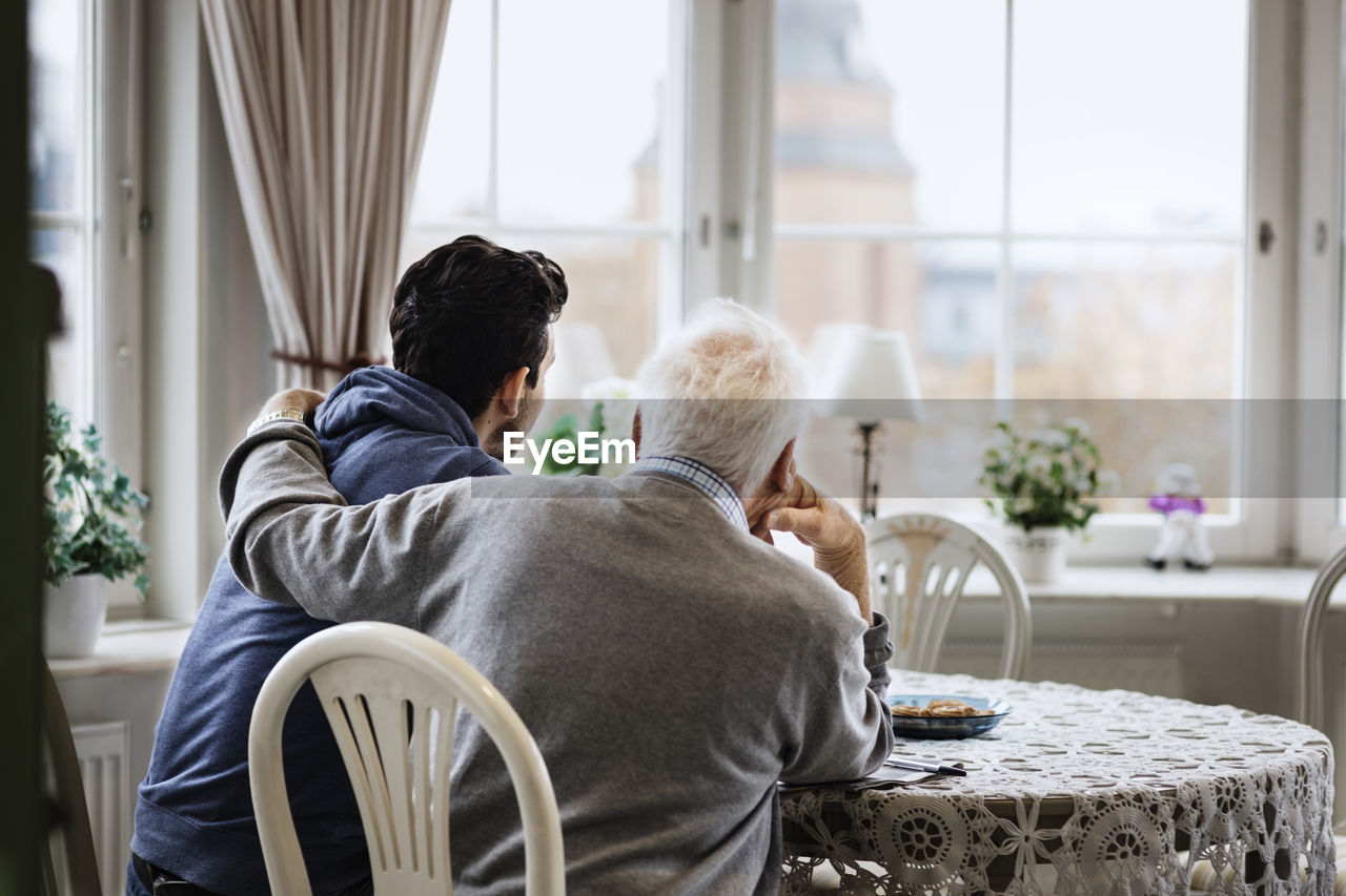Rear view of man sitting with arm around caretaker at nursing home