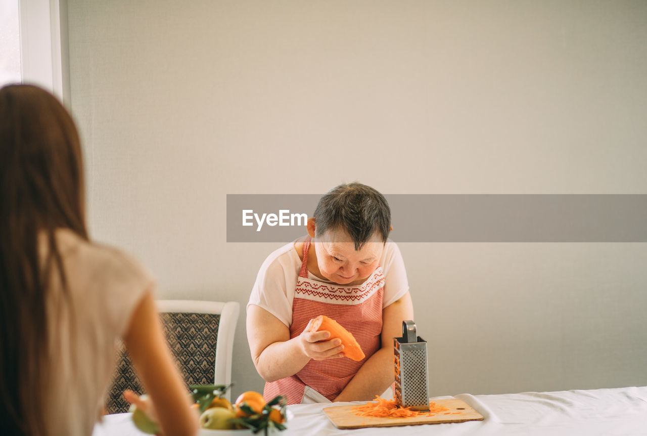 Lifestyle, education. an elderly woman with down syndrome rubs carrots on a grater with an assistant