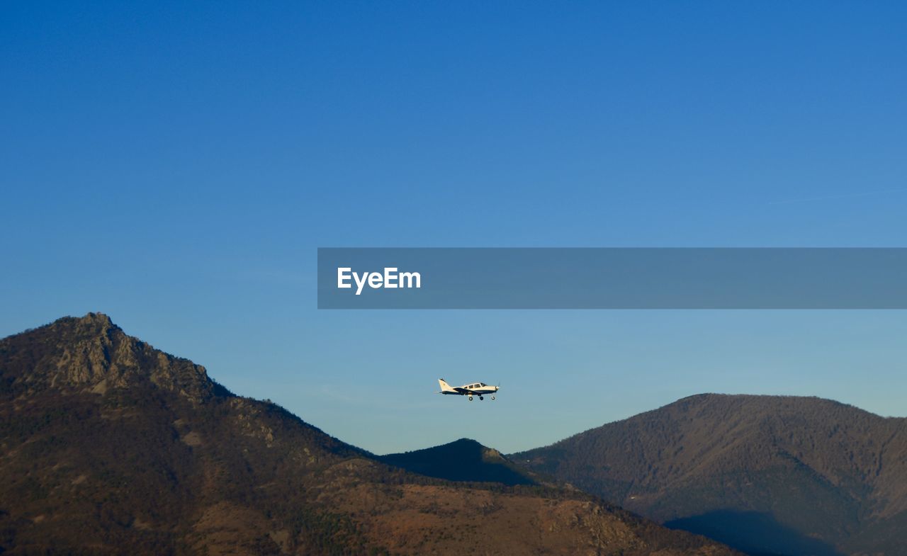 Low angle view of airplane flying over mountains against blue sky