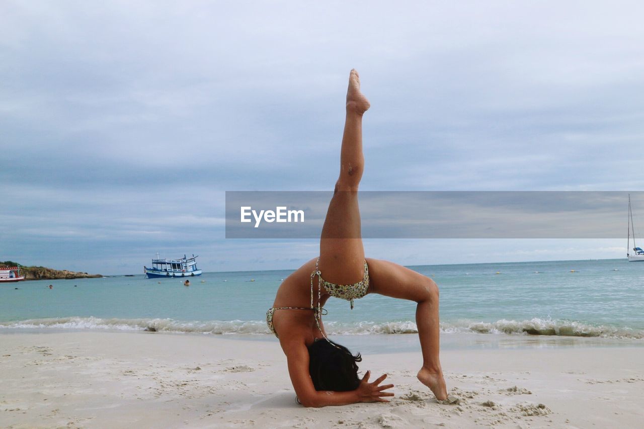 Full length of young woman doing headstand at beach against cloudy sky